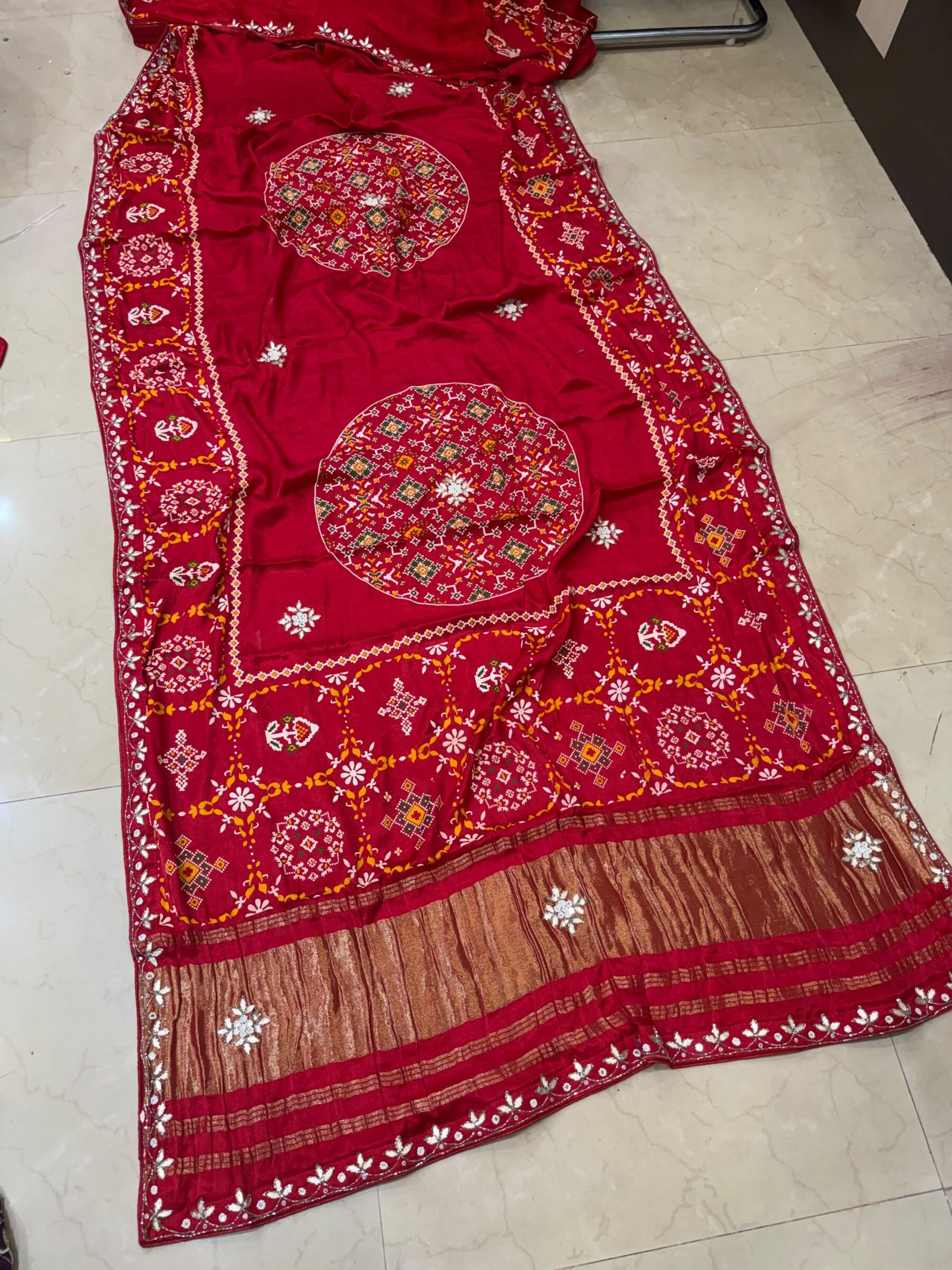 Pure Modal Gajji Patola Saree with Gota Patti Work 10 A beautiful red pure Modal Gajji Patola saree with intricate Gota Patti handwork and a metallic zari border spread across a tiled floor.