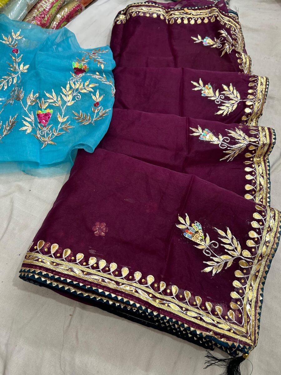A beautiful deep purple Organza saree with detailed gold Gota patti handwork and a contrasting bright blue blouse piece.