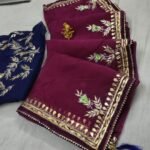 A beautiful deep purple Organza saree with detailed gold Gota patti handwork and a contrasting bright blue blouse piece.