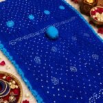 A beautiful bright blue Jodhpuri Bandhani saree made of Nazneen chiffon displayed with traditional decor, flowers, and bangles.