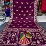 A wine coloured Dola silk saree featuring mirror work, zari butti, and an embroidered border laid out flat.