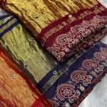 A close-up of a beautifully folded Modal Gajji Silk saree featuring rich golden and red hues with traditional mirror work and intricate hand-print patterns.