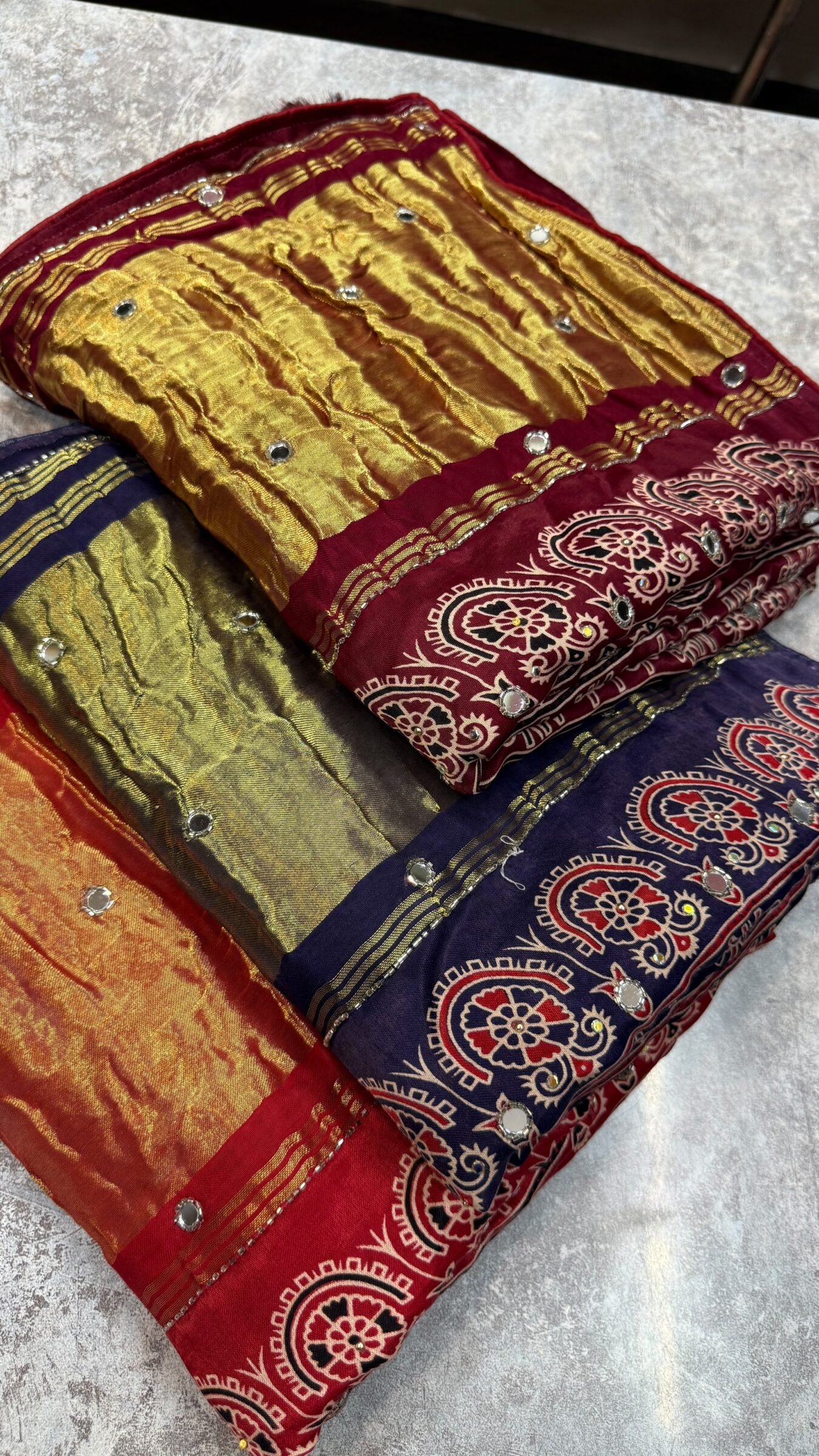 Pure Modal Gajji Silk Mirror Work Saree 1 A close-up of a beautifully folded Modal Gajji Silk saree featuring rich golden and red hues with traditional mirror work and intricate hand-print patterns.