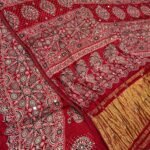 A close-up of a beautifully folded Modal Gajji Silk saree featuring rich golden and red hues with traditional mirror work and intricate hand-print patterns.