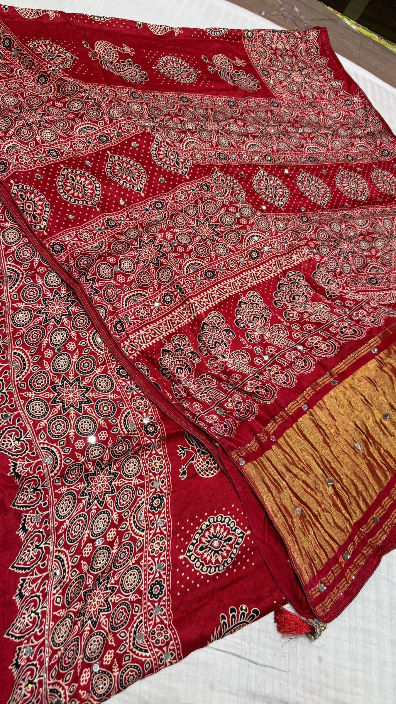 Pure Modal Gajji Silk Mirror Work Saree 3 A close-up of a beautifully folded Modal Gajji Silk saree featuring rich golden and red hues with traditional mirror work and intricate hand-print patterns.