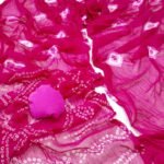 A vibrant red Nazneen chiffon saree with Bandhani print and a matching pink blouse piece spread out on a white surface.
