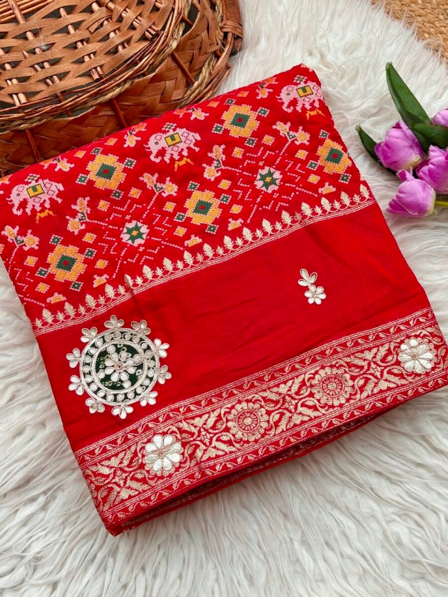 A vibrant red Patola print saree with gota patti work folded on a white faux fur rug next to a basket and purple tulips.