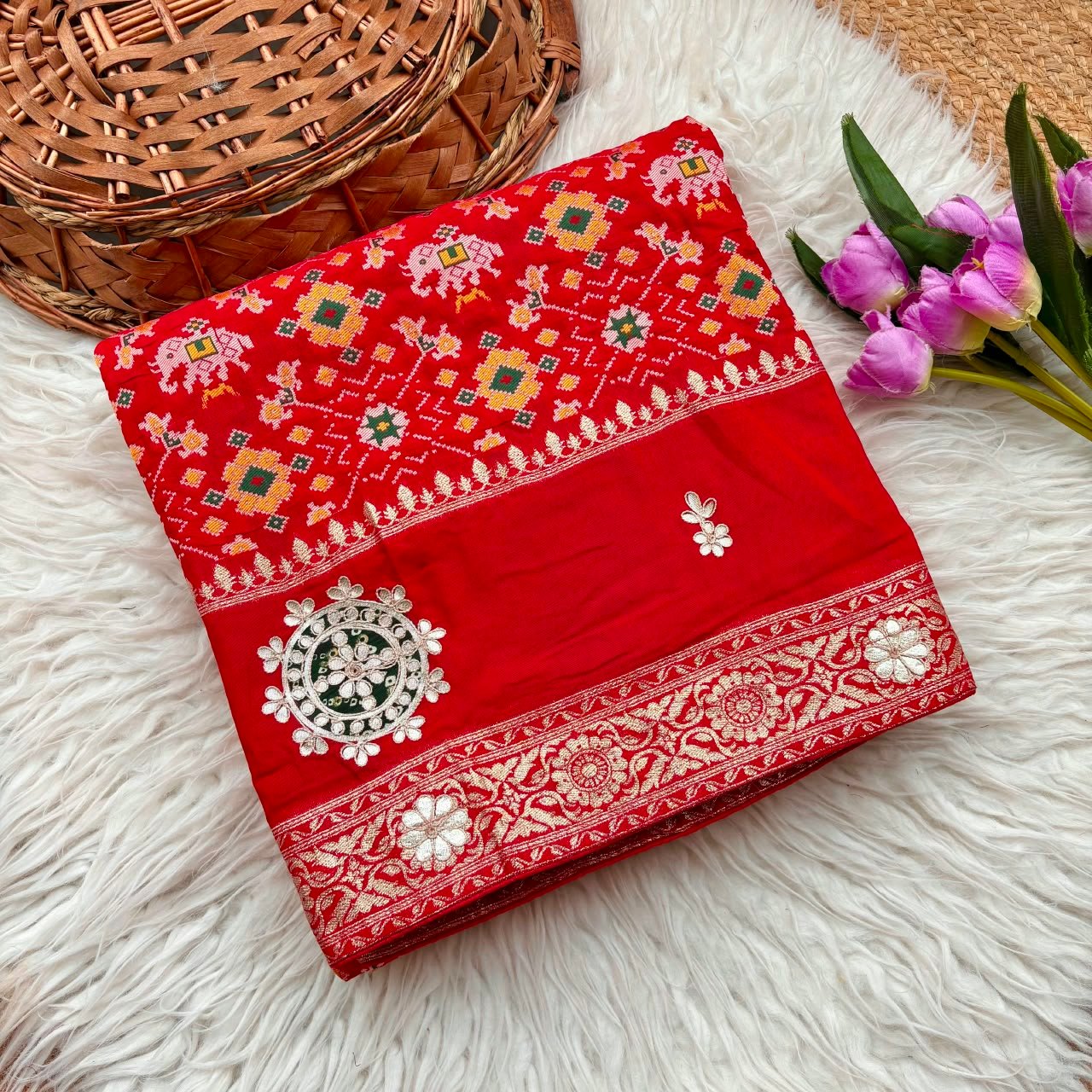 Patola Saree with Gota Patti Work on Viscose Dolla Silk 1 A vibrant red Patola print saree with gota patti work folded on a white faux fur rug next to a basket and purple tulips.