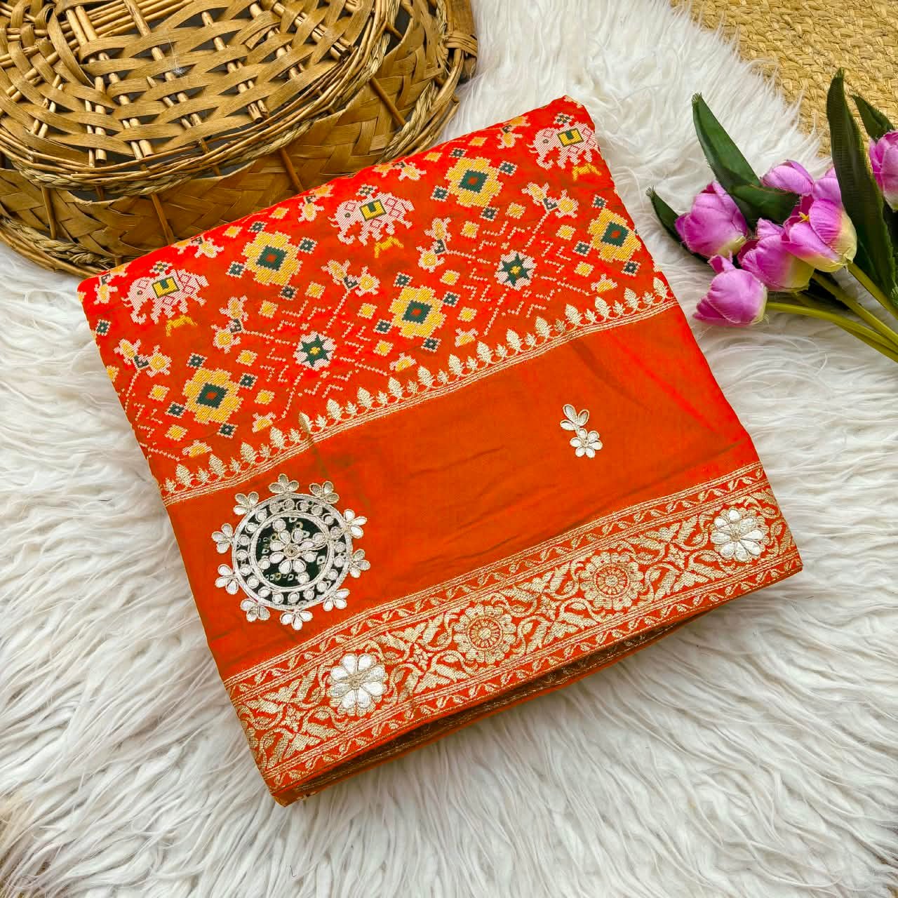Patola Saree with Gota Patti Work on Viscose Dolla Silk 2 A vibrant red Patola print saree with gota patti work folded on a white faux fur rug next to a basket and purple tulips.