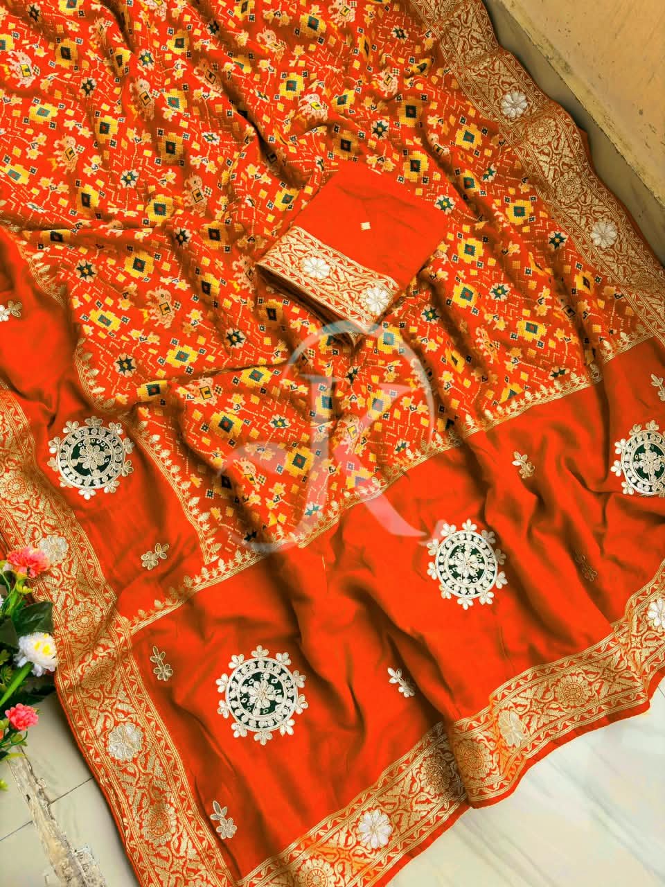 Patola Saree with Gota Patti Work on Viscose Dolla Silk 3 A vibrant red Patola print saree with gota patti work folded on a white faux fur rug next to a basket and purple tulips.