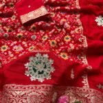 A vibrant red Patola print saree with gota patti work folded on a white faux fur rug next to a basket and purple tulips.