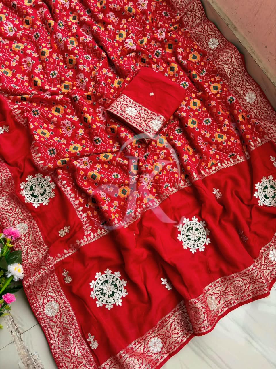 Patola Saree with Gota Patti Work on Viscose Dolla Silk 5 A vibrant red Patola print saree with gota patti work folded on a white faux fur rug next to a basket and purple tulips.