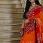 A woman wearing a vibrant orange and red Piliya saree with Gota Patti work standing on a staircase.