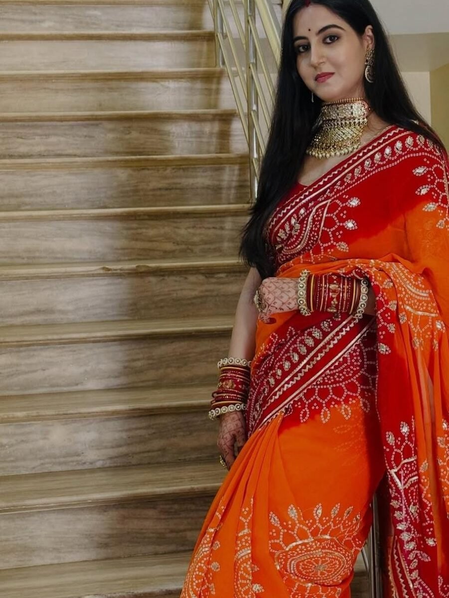 A woman wearing a vibrant orange and red Piliya saree with Gota Patti work standing on a staircase.