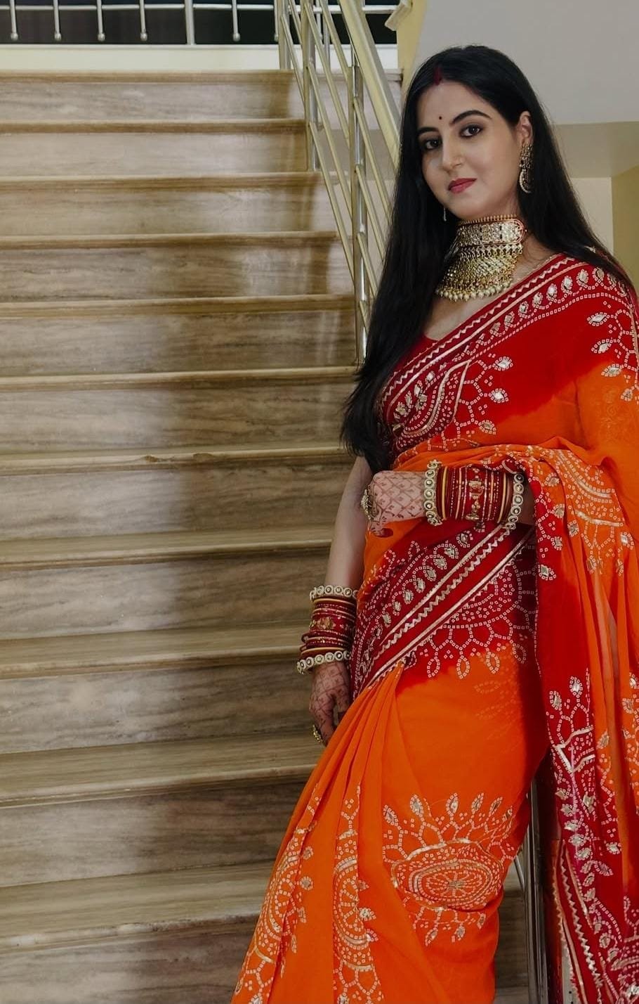Piliya Saree in Georgette with Gota Patti Work 1 A woman wearing a vibrant orange and red Piliya saree with Gota Patti work standing on a staircase.