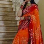 A woman wearing a vibrant orange and red Piliya saree with Gota Patti work standing on a staircase.