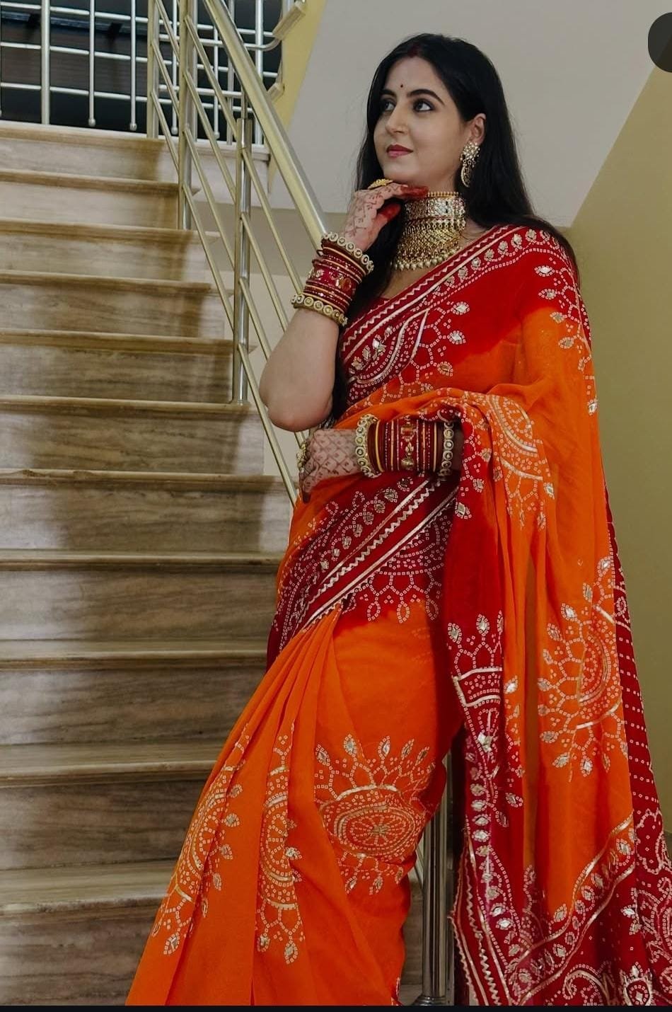Piliya Saree in Georgette with Gota Patti Work 2 A woman wearing a vibrant orange and red Piliya saree with Gota Patti work standing on a staircase.