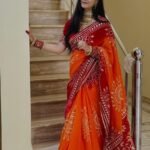 A woman wearing a vibrant orange and red Piliya saree with Gota Patti work standing on a staircase.