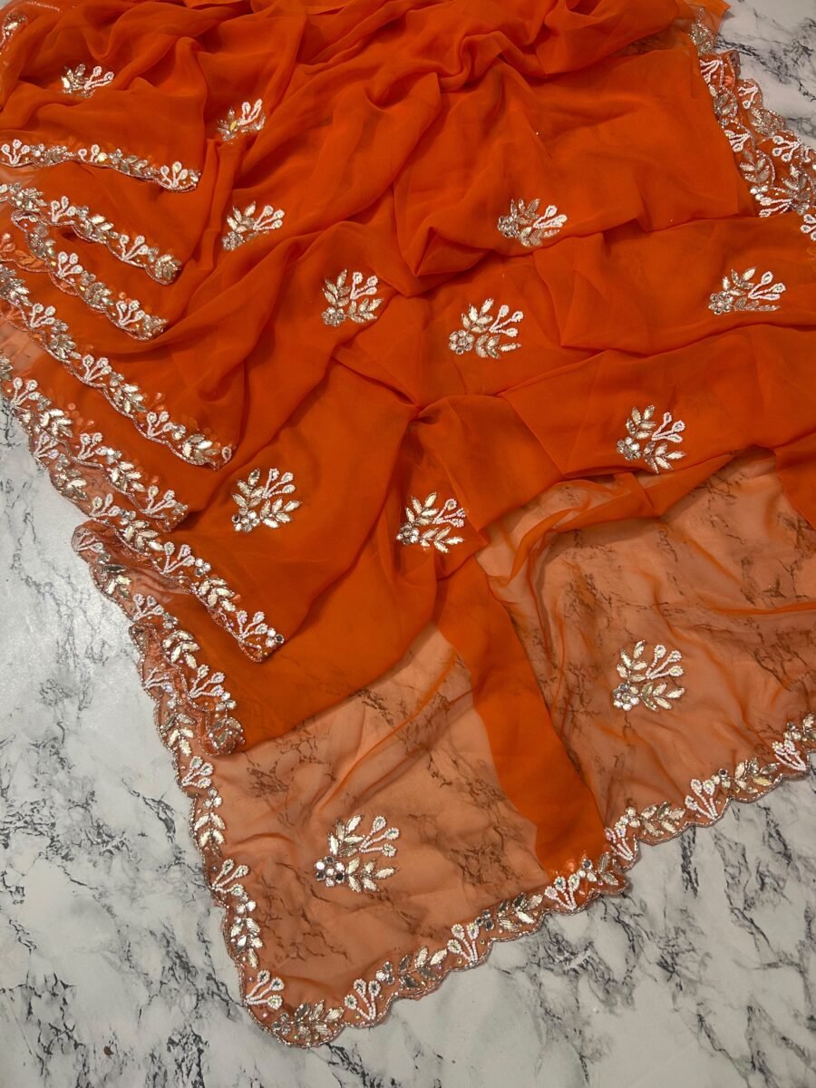 A vibrant orange Royal Georgette saree laid flat, featuring intricate white handwork floral motifs and a detailed cutwork border.