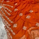 A vibrant orange Royal Georgette saree laid flat, featuring intricate white handwork floral motifs and a detailed cutwork border.
