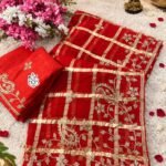 A luxurious red soft viscose silk saree with gold grid patterns and intricate floral embroidery border displayed on a white fluffy rug.