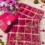 A luxurious red soft viscose silk saree with gold grid patterns and intricate floral embroidery border displayed on a white fluffy rug.