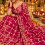 A luxurious red soft viscose silk saree with gold grid patterns and intricate floral embroidery border displayed on a white fluffy rug.