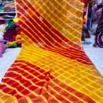 A vibrant Tora Silk Leheriya saree featuring diagonal yellow, pink, and dark green stripes laid out on a white surface.