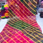 A vibrant Tora Silk Leheriya saree featuring diagonal yellow, pink, and dark green stripes laid out on a white surface.